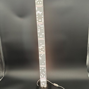 May include: A tall, gray skyscraper lamp with a black base. The lamp is illuminated with white and pink lights, resembling windows. A ruler is placed in front of the lamp. The lamp has a white cord and a switch.