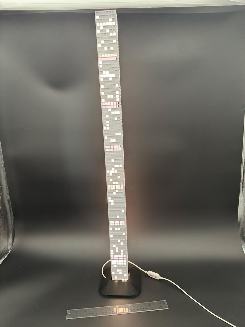 May include: A tall, gray skyscraper lamp with a black base. The lamp is illuminated with white and pink lights, resembling windows. A ruler is placed in front of the lamp. The lamp has a white cord and a switch.