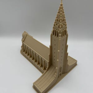 May include: A tan-colored architectural model of a church with a tall, pointed steeple and a cross at the top. The model has detailed features, including arched windows and vertical lines, and is set against a plain background.