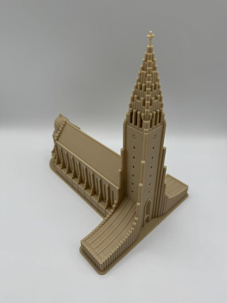 May include: A tan-colored architectural model of a church with a tall, pointed steeple and a cross at the top. The model has detailed features, including arched windows and vertical lines, and is set against a plain background.