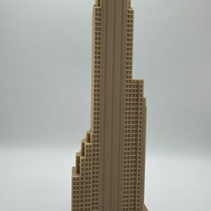 May include: A tan-colored miniature skyscraper model with a detailed facade featuring numerous windows. The building has a stepped design and a tall, central tower. The model is likely made of plastic or a similar material.