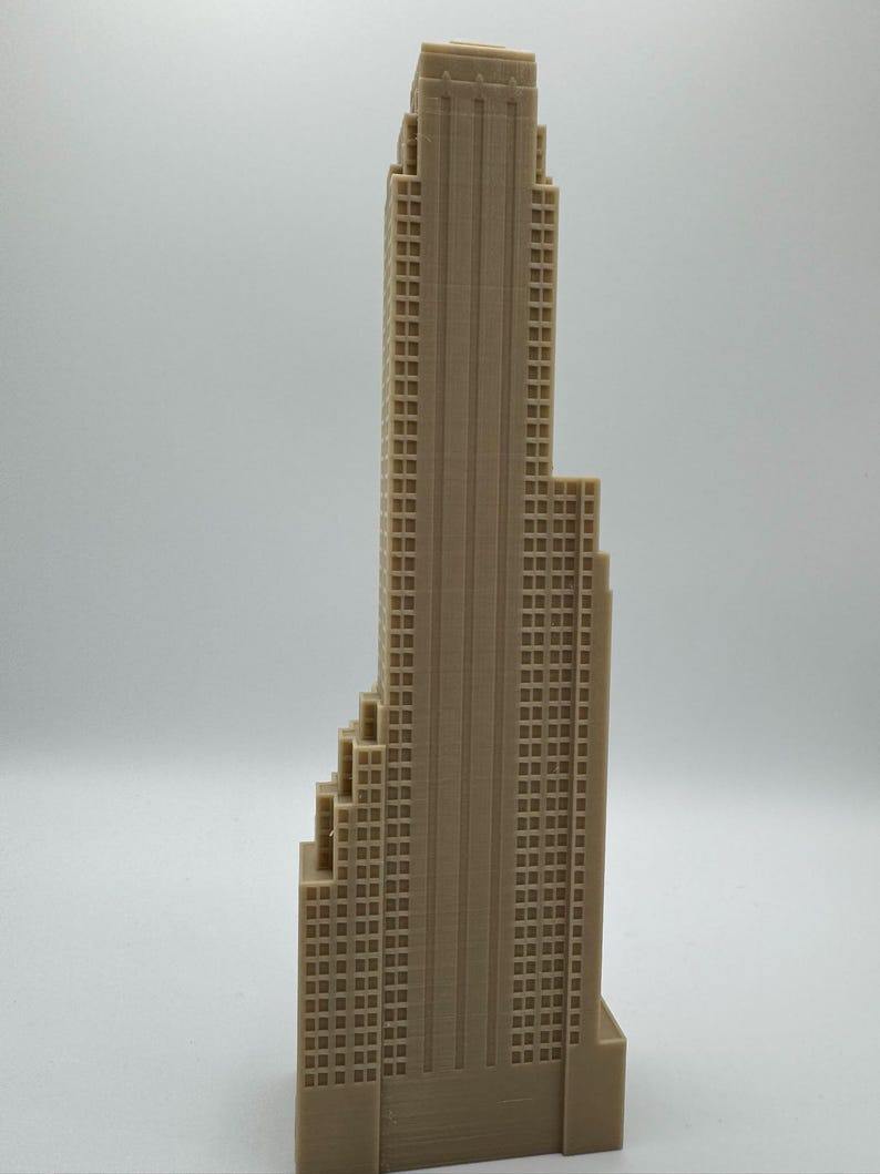May include: A tan-colored miniature skyscraper model with a detailed facade featuring numerous windows. The building has a stepped design and a tall, central tower. The model is likely made of plastic or a similar material.