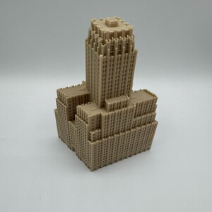 May include: A tan-colored miniature model of a skyscraper. The building has a tiered design with multiple rectangular sections and numerous small windows. The model is made of a solid material and sits on a white surface.
