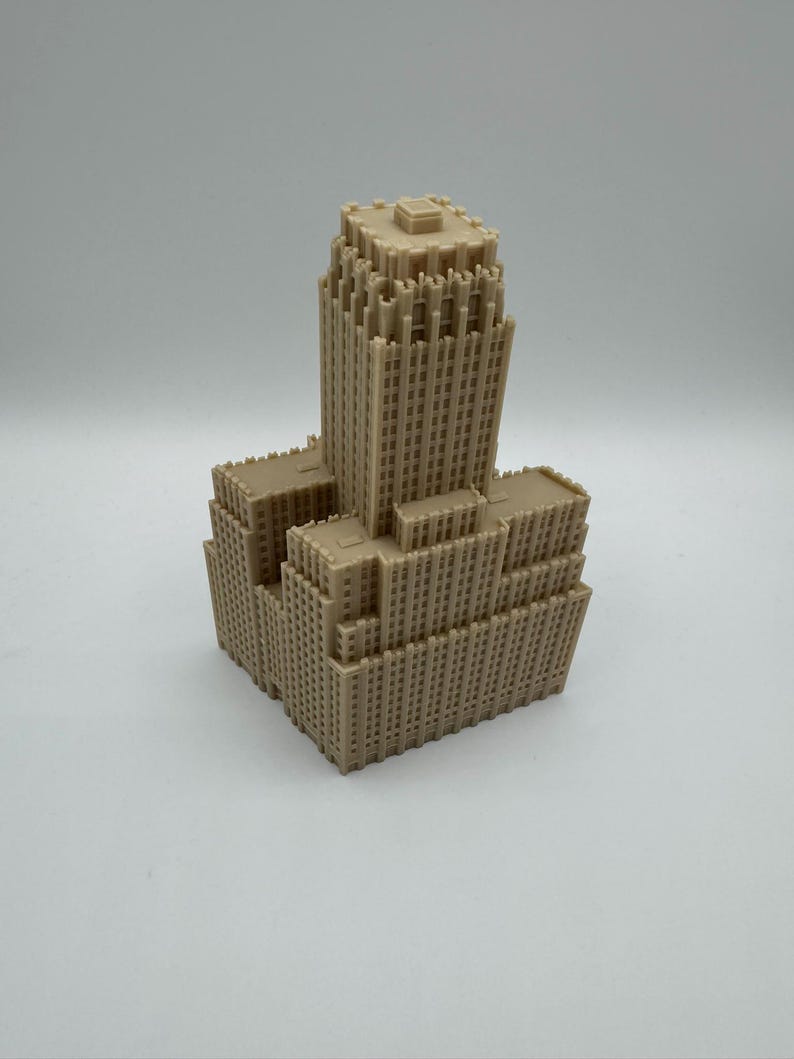 May include: A tan-colored miniature model of a skyscraper. The building has a tiered design with multiple rectangular sections and numerous small windows. The model is made of a solid material and sits on a white surface.