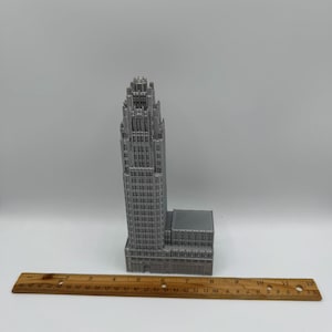 May include: A gray, 3D-printed model of a skyscraper with intricate architectural details. The building is tall and slender, with a detailed top. A wooden ruler is placed in front of the building. The building is approximately 10 inches tall.