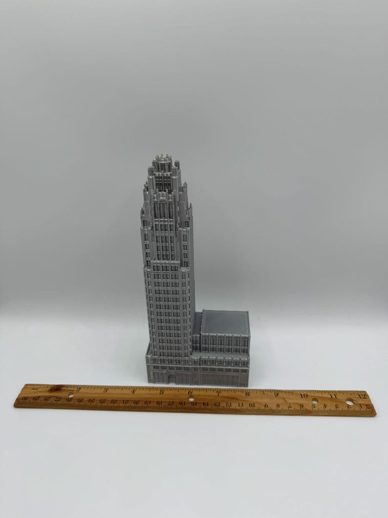 May include: A gray, 3D-printed model of a skyscraper with intricate architectural details. The building is tall and slender, with a detailed top. A wooden ruler is placed in front of the building. The building is approximately 10 inches tall.