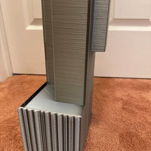 K&ouml;nnte beinhalten: Ein graues, 3D-gedrucktes Architekturmodell mit strukturierter Oberfl&auml;che. Das Modell hat eine rechteckige Basis mit vertikalen Rippen und eine turmartige Struktur. Das Design umfasst einen ausgesparten Bereich und eine horizontale Erweiterung.