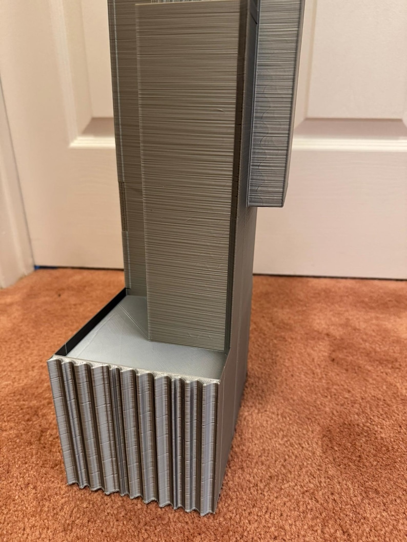 K&ouml;nnte beinhalten: Ein graues, 3D-gedrucktes Architekturmodell mit strukturierter Oberfl&auml;che. Das Modell hat eine rechteckige Basis mit vertikalen Rippen und eine turmartige Struktur. Das Design umfasst einen ausgesparten Bereich und eine horizontale Erweiterung.