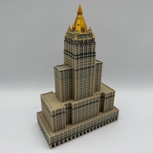 May include: A detailed model of a skyscraper with a tan facade and a gold-colored spire. The building has many windows and architectural details, showcasing a classic design. The model is a miniature representation of a famous landmark.