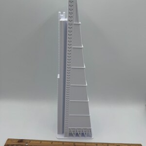 May include: A white architectural model of a tall building with a triangular shape. The model has detailed features, including a ladder-like structure and a series of horizontal lines. A wooden ruler is placed at the bottom, showing measurements in inches.