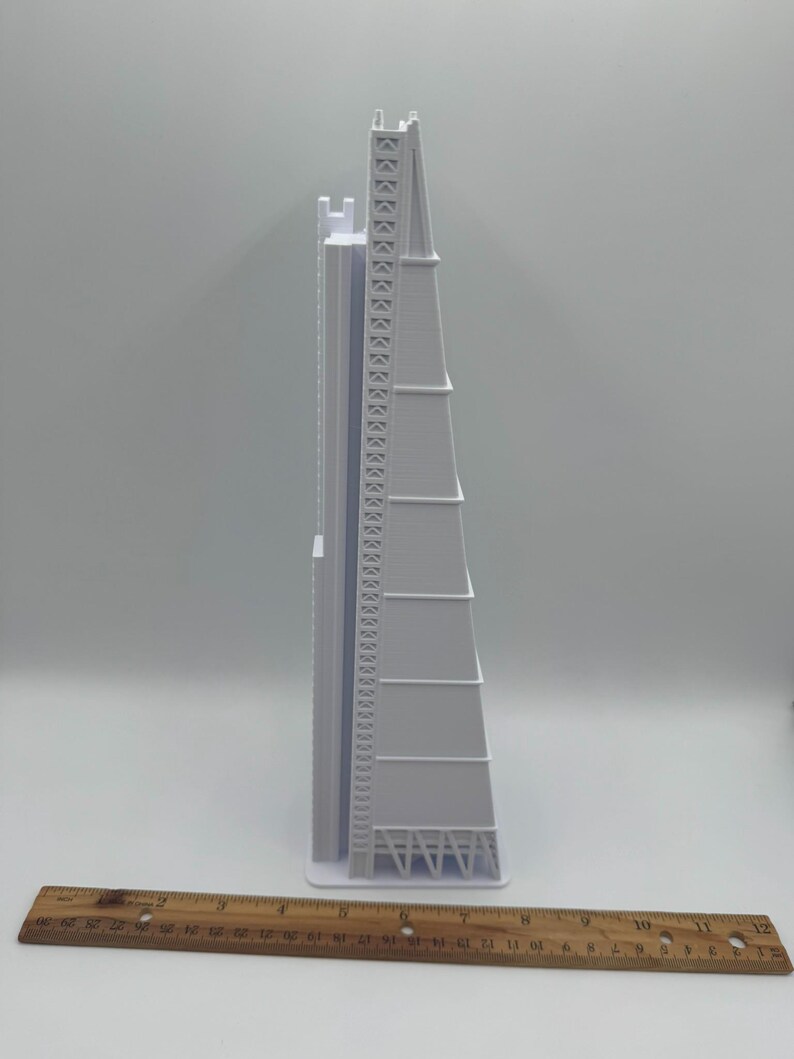 May include: A white architectural model of a tall building with a triangular shape. The model has detailed features, including a ladder-like structure and a series of horizontal lines. A wooden ruler is placed at the bottom, showing measurements in inches.