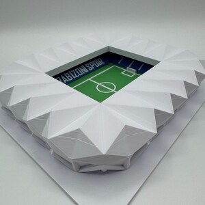May include: A white, geometric model of a soccer stadium. The stadium has a green field with white markings and the text "Trabzonspor" in blue. The stands are white and angled, creating a detailed architectural design.