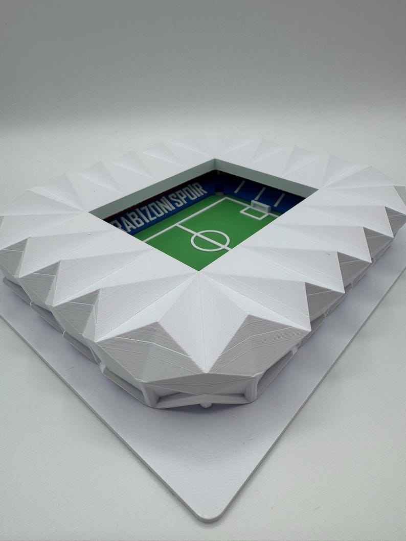 May include: A white, geometric model of a soccer stadium. The stadium has a green field with white markings and the text "Trabzonspor" in blue. The stands are white and angled, creating a detailed architectural design.