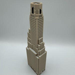May include: A beige 3D printed architectural model of a skyscraper featuring a stepped design with multiple levels, a grid of small square windows, and a flat roof section. The structure has a vertical orientation with a central tower section.