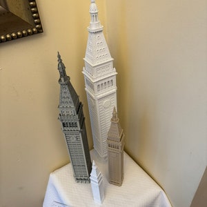 May include: Four miniature white, gray, and brown buildings with pointed roofs. The buildings are arranged in a triangular formation on a white tablecloth. A ruler is visible in the bottom left corner of the image.