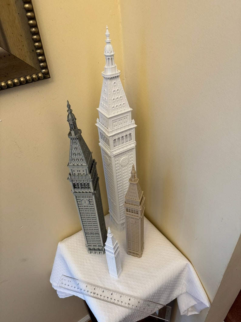 May include: Four miniature white, gray, and brown buildings with pointed roofs. The buildings are arranged in a triangular formation on a white tablecloth. A ruler is visible in the bottom left corner of the image.