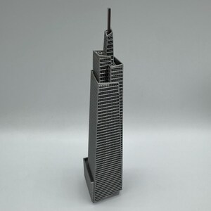 May include: A miniature gray skyscraper model with a detailed facade and a tall antenna. The building has a rectangular base and a multi-tiered design, showcasing architectural detail. The model is set against a plain background.