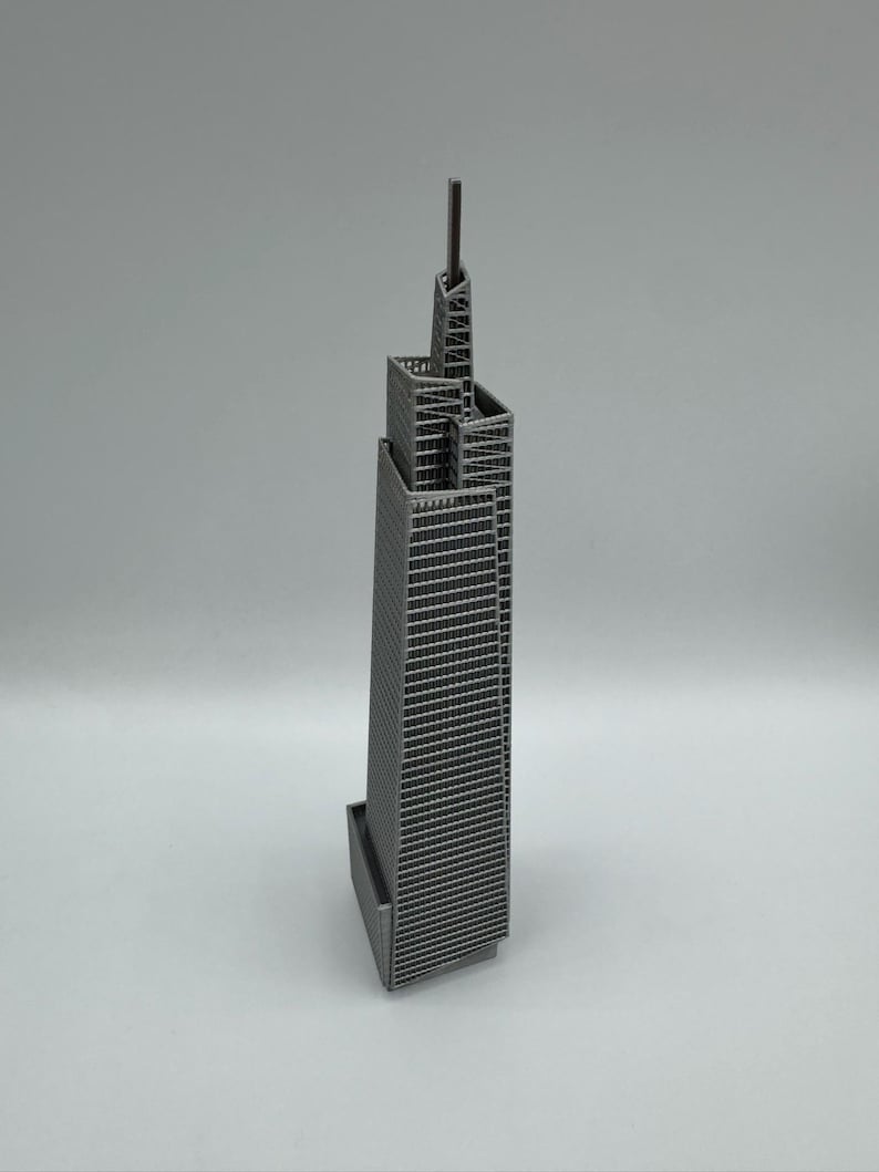 May include: A miniature gray skyscraper model with a detailed facade and a tall antenna. The building has a rectangular base and a multi-tiered design, showcasing architectural detail. The model is set against a plain background.