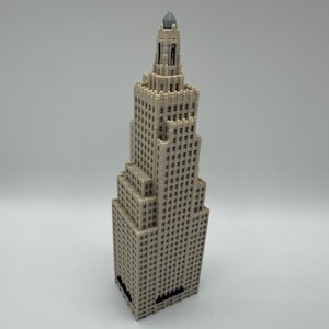 May include: A beige miniature skyscraper model with a detailed facade, featuring numerous windows and a tiered design. The model has a pointed top and a dark base, set against a plain background.