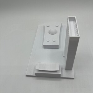 K&ouml;nnte beinhalten: Wei&szlig;es 3D-gedrucktes Modell eines Geb&auml;udes mit einem Kuppeldach, einem rechteckigen Geb&auml;ude mit einem Balkon und einer geschwungenen Bank. Das Modell steht auf einem wei&szlig;en Sockel.