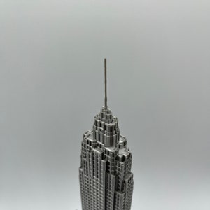 K&ouml;nnte beinhalten: Eine detaillierte, silberfarbene Miniatur des Empire State Building. Das Modell weist komplizierte architektonische Details auf, einschlie&szlig;lich der Antenne und mehrerer Ebenen. Das Geb&auml;ude steht vor einem schlichten, hellen Hintergrund.