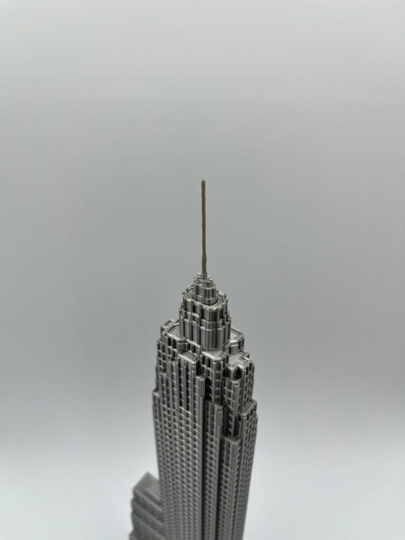 K&ouml;nnte beinhalten: Eine detaillierte, silberfarbene Miniatur des Empire State Building. Das Modell weist komplizierte architektonische Details auf, einschlie&szlig;lich der Antenne und mehrerer Ebenen. Das Geb&auml;ude steht vor einem schlichten, hellen Hintergrund.