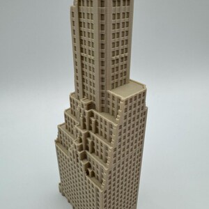 May include: A beige, miniature skyscraper model with a detailed facade featuring numerous windows. The building has a stepped design, with a tall central tower and multiple lower sections. The model is likely made of plastic or a similar material.