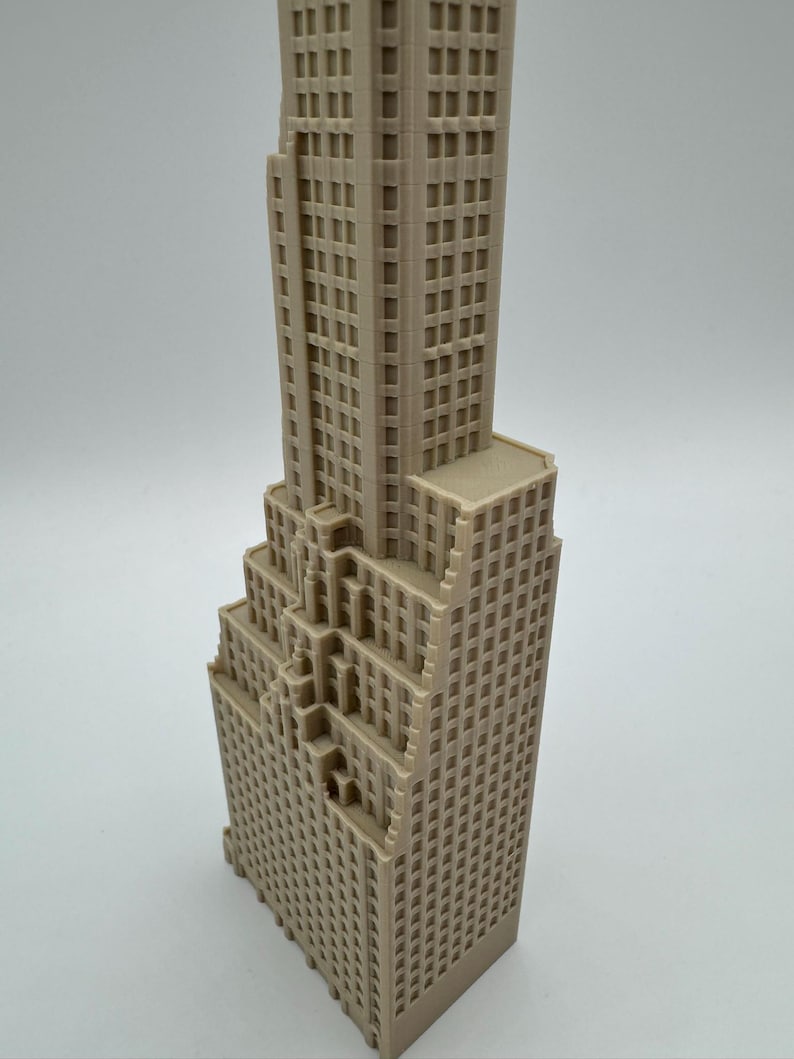 May include: A beige, miniature skyscraper model with a detailed facade featuring numerous windows. The building has a stepped design, with a tall central tower and multiple lower sections. The model is likely made of plastic or a similar material.