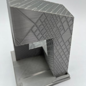 Peut inclure: Mod&egrave;le d'immeuble imprim&eacute; en 3D de couleur grise avec un design g&eacute;om&eacute;trique. Le mod&egrave;le a une base carr&eacute;e et une structure rectangulaire haute avec un toit angulaire unique.