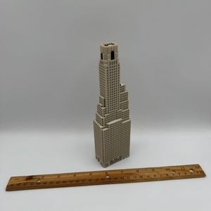 May include: A beige 3D printed model of a skyscraper with a stepped architectural design, positioned next to a wooden ruler that shows the model stands approximately 19 cm tall. The structure features a grid of small square windows.