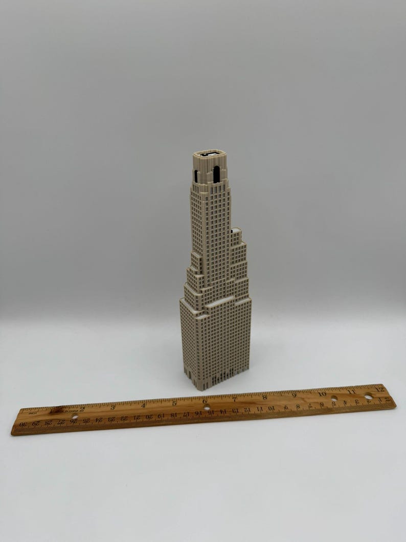 May include: A beige 3D printed model of a skyscraper with a stepped architectural design, positioned next to a wooden ruler that shows the model stands approximately 19 cm tall. The structure features a grid of small square windows.