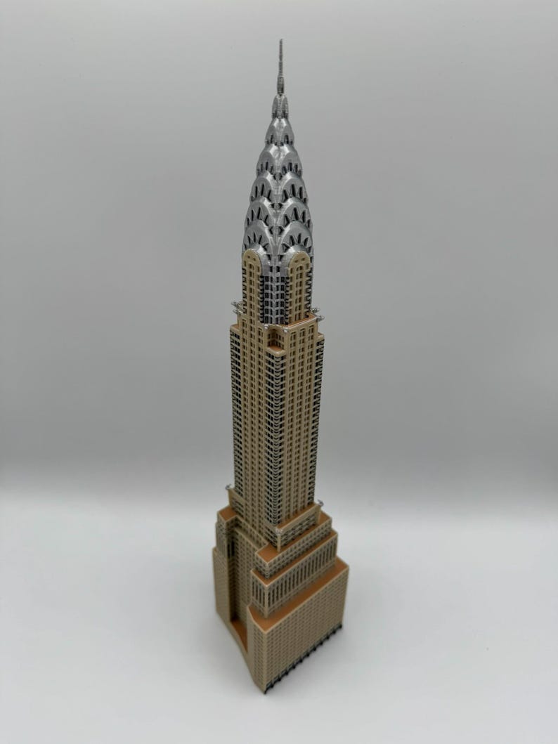 May include: A detailed model of the Chrysler Building, a famous Art Deco skyscraper. The model features a silver spire and a tan and brown facade, showcasing the building's distinctive architectural details. The model is a miniature replica of the iconic New York City landmark.