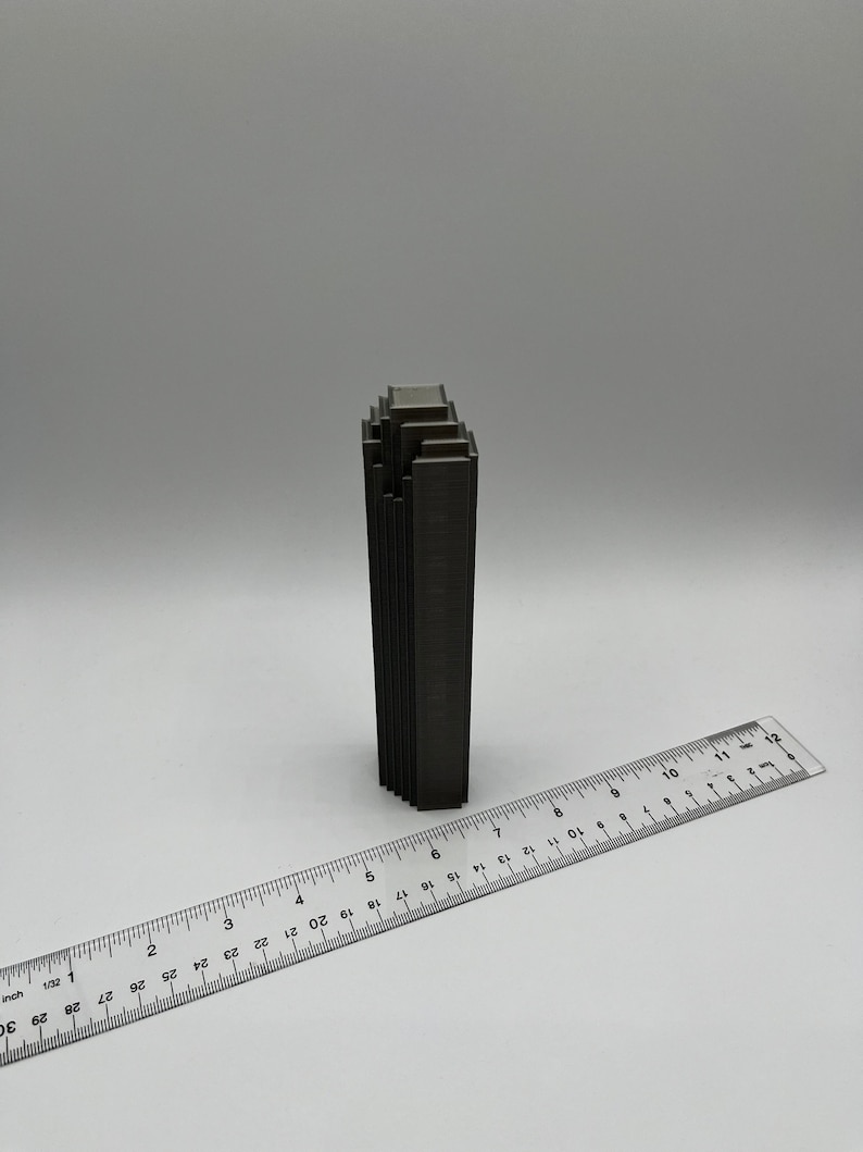 May include: A dark gray metal sculpture of a building with a stacked design. The sculpture is about 6 inches tall and is sitting on a white surface.