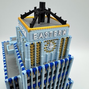 May include: A 3D-printed model of a building with the word "EASTERN" in white letters. The building features a light blue facade with gold and blue accents. The top has a black structure.