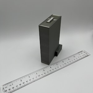 May include: A gray, detailed model of a skyscraper building with a flat roof. The model is approximately 7 inches tall and is sitting on a white surface next to a ruler.