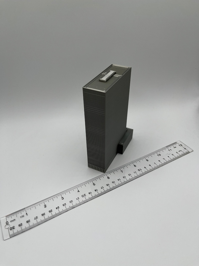 May include: A gray, detailed model of a skyscraper building with a flat roof. The model is approximately 7 inches tall and is sitting on a white surface next to a ruler.