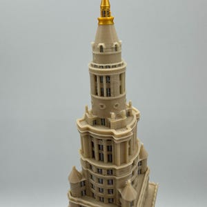 May include: A beige model of a tall building with a gold spire. The building has multiple levels with windows and architectural details. The spire is a bright gold color, and the building is a light beige.