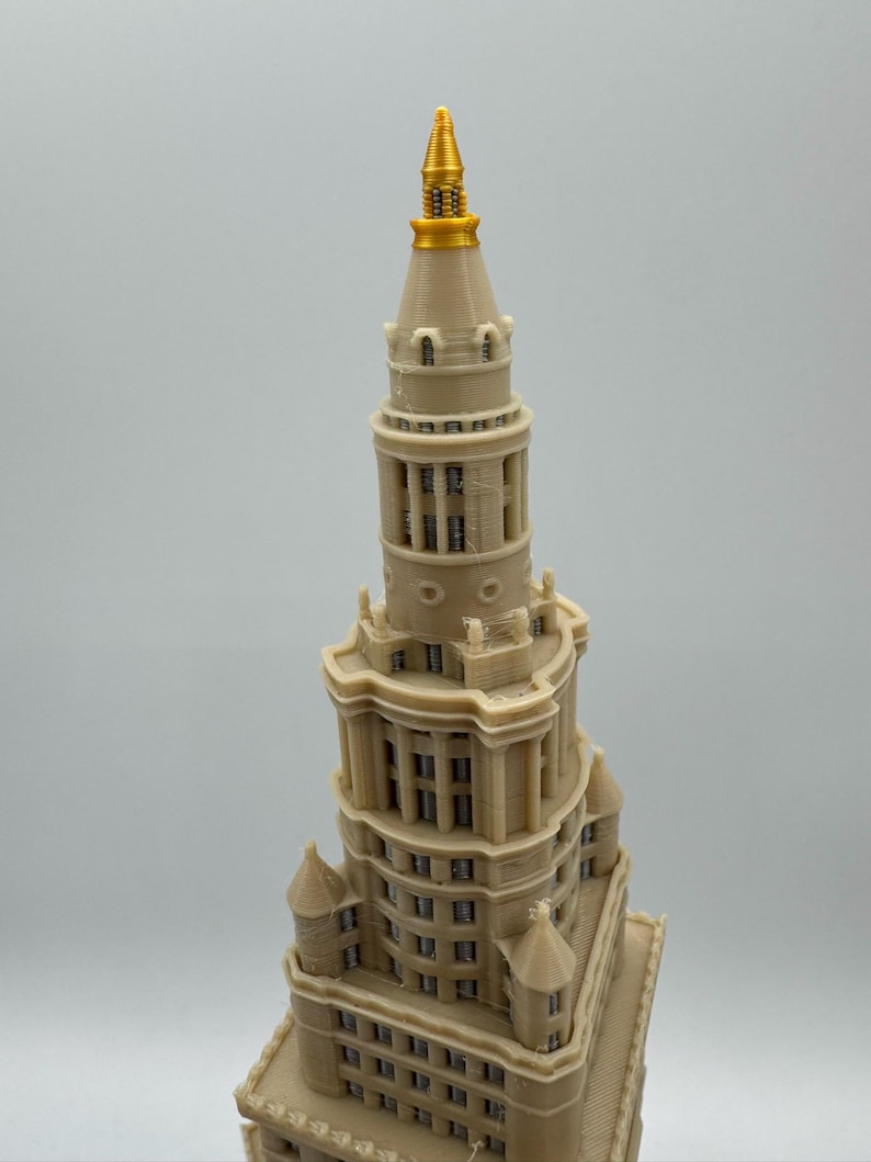 May include: A beige model of a tall building with a gold spire. The building has multiple levels with windows and architectural details. The spire is a bright gold color, and the building is a light beige.