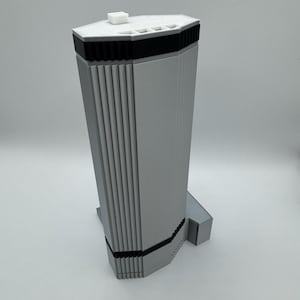 May include: A silver-colored, geometric knife block with a white top and black accents. The knife block has a rectangular base and a tall, multi-sided body with vertical grooves. A small, white square is on top.