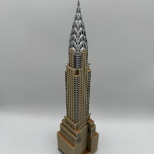 May include: A detailed miniature of the Chrysler Building, a famous skyscraper. The model features a tan base and body with a silver-colored crown and spire. The building's architectural details are clearly visible.