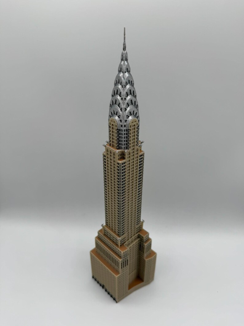 May include: A detailed miniature of the Chrysler Building, a famous skyscraper. The model features a tan base and body with a silver-colored crown and spire. The building's architectural details are clearly visible.