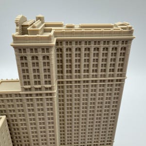 May include: A detailed, beige-colored architectural model of a skyscraper. The model features numerous windows, vertical columns, and intricate rooftop details. The building's design showcases a classic, historical style.