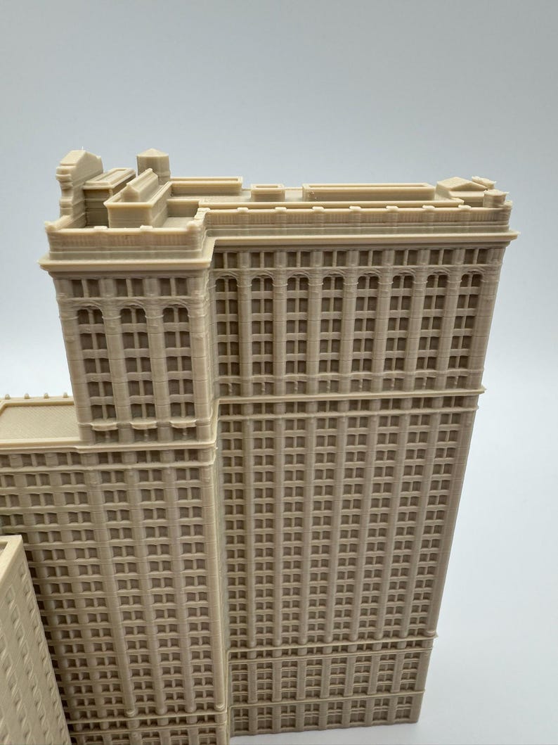 May include: A detailed, beige-colored architectural model of a skyscraper. The model features numerous windows, vertical columns, and intricate rooftop details. The building's design showcases a classic, historical style.
