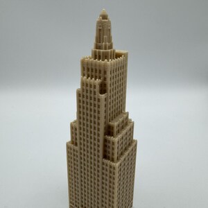 May include: A tan-colored miniature skyscraper model with a detailed, multi-tiered design. The building features numerous windows and architectural details, showcasing a classic skyscraper aesthetic. The model is a solid, three-dimensional object.