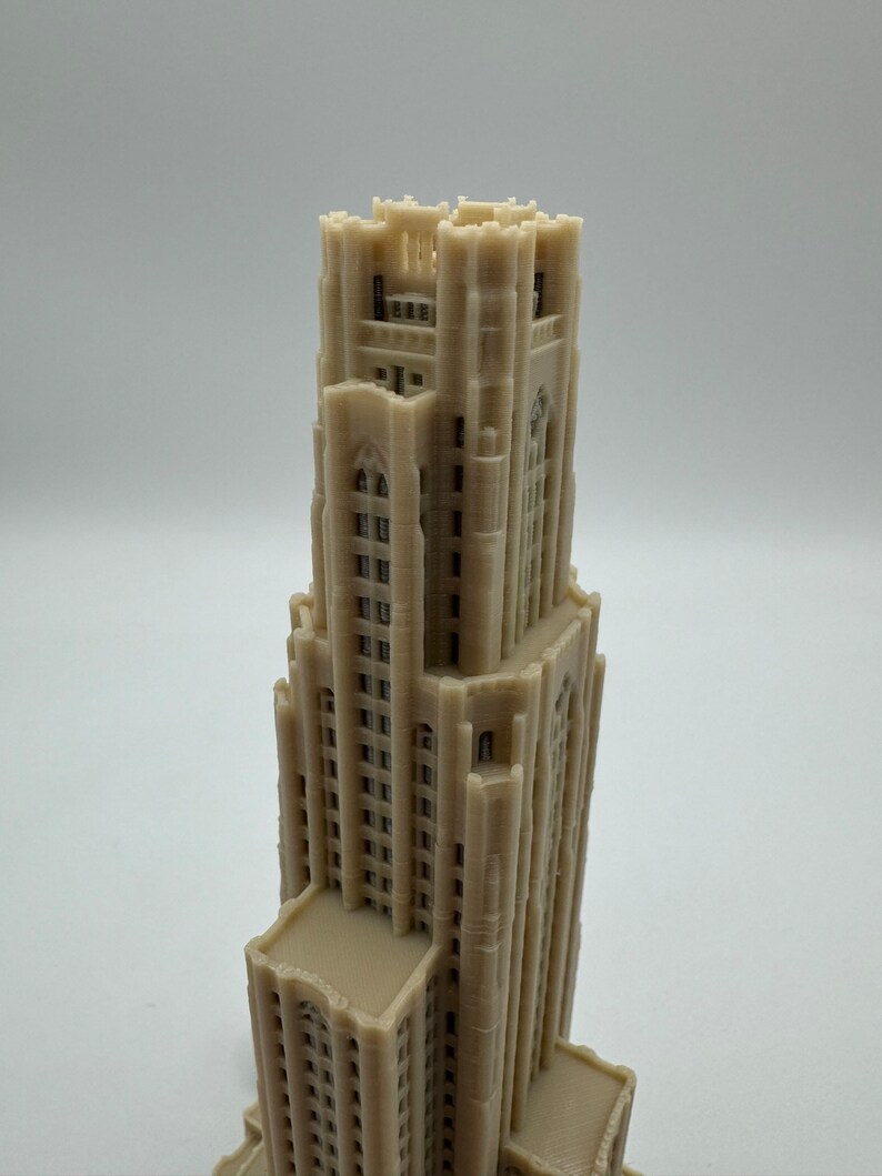 May include: A 3D printed model of a tall, beige building with a pointed roof and many windows. The building has a detailed facade with intricate carvings and decorative elements.