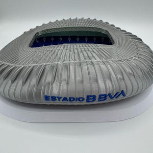May include: A detailed, gray stadium model with the text "ESTADIO BBVA" in blue. The model has a textured exterior and an open interior with blue accents. The stadium is designed with intricate architectural details.