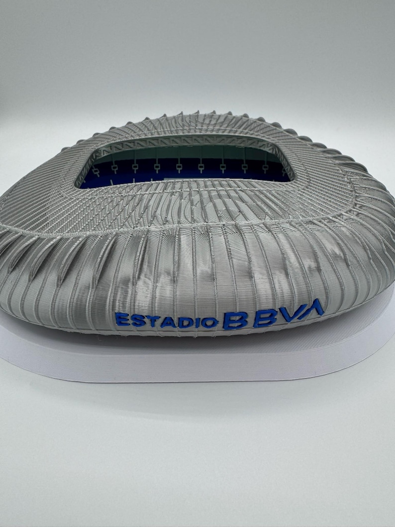 May include: A detailed, gray stadium model with the text "ESTADIO BBVA" in blue. The model has a textured exterior and an open interior with blue accents. The stadium is designed with intricate architectural details.