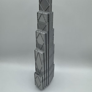 May include: A gray 3D printed model of a skyscraper featuring a stepped, tiered design. The structure displays a prominent exterior cross-bracing pattern on its facade, with vertical lines and rectangular segments forming the building's frame.