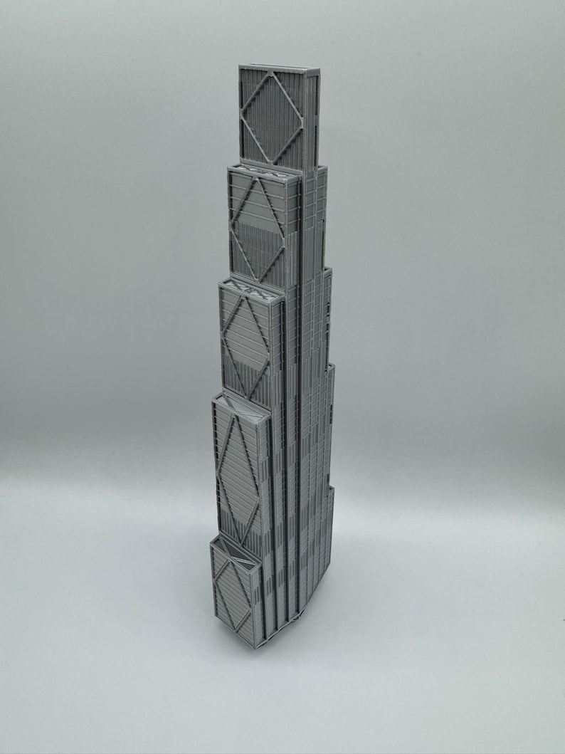 May include: A gray 3D printed model of a skyscraper featuring a stepped, tiered design. The structure displays a prominent exterior cross-bracing pattern on its facade, with vertical lines and rectangular segments forming the building's frame.