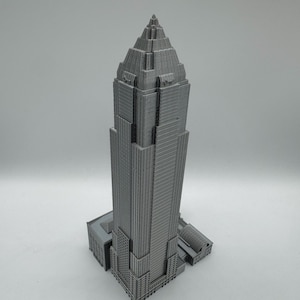 May include: A gray, 3D-printed model of a skyscraper. The building has a tiered design with a pointed top and detailed architectural features. The model is set against a plain white background.